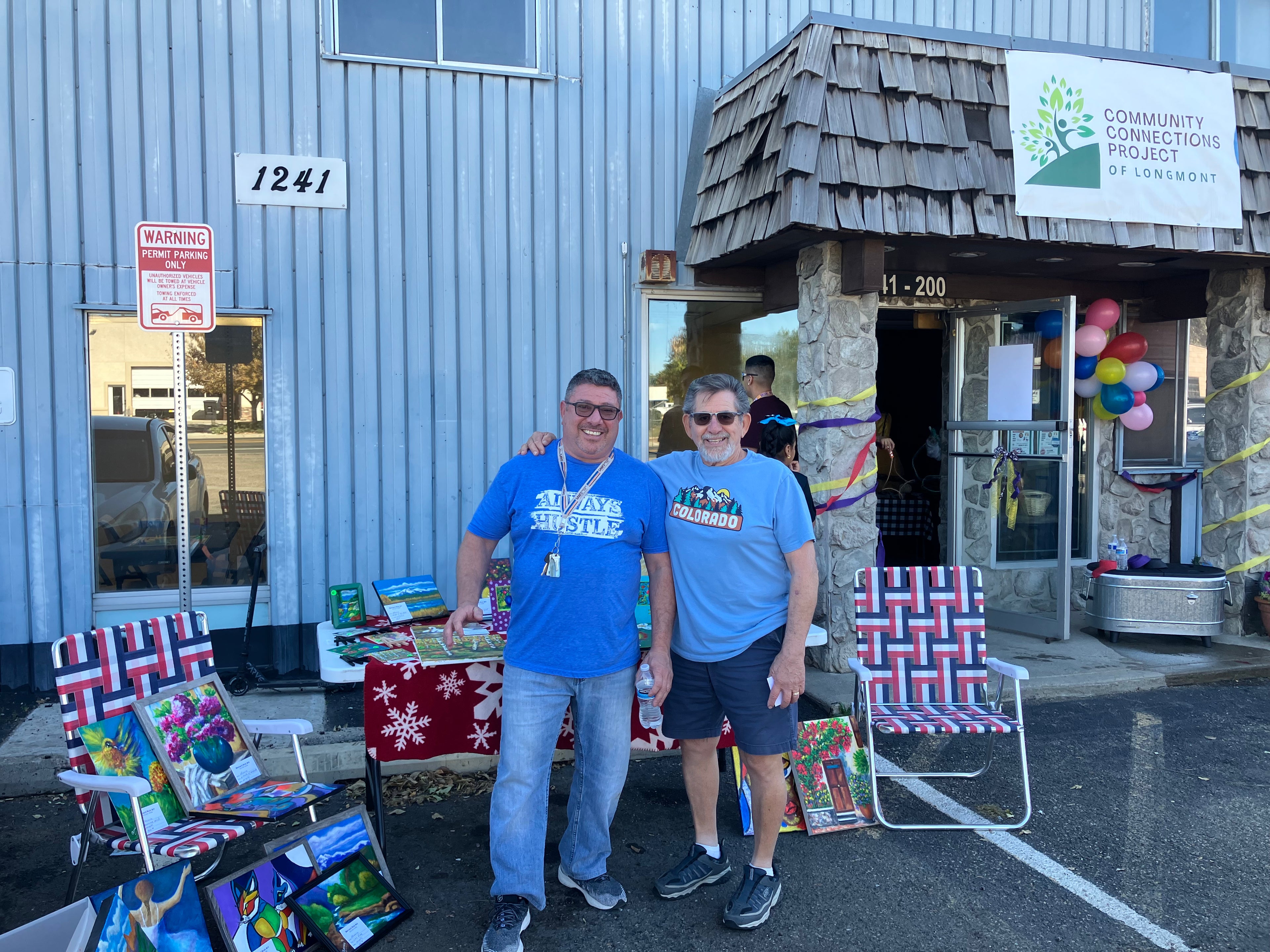 2 people outside of commercial space with a table of art for sale and balloons in the background